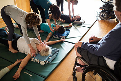 Ambulatory Adaptive Yoga. Several people lying with arms spread wide on a green gym mat. Assistants standing above them, placing gentle reference at their shoulders.