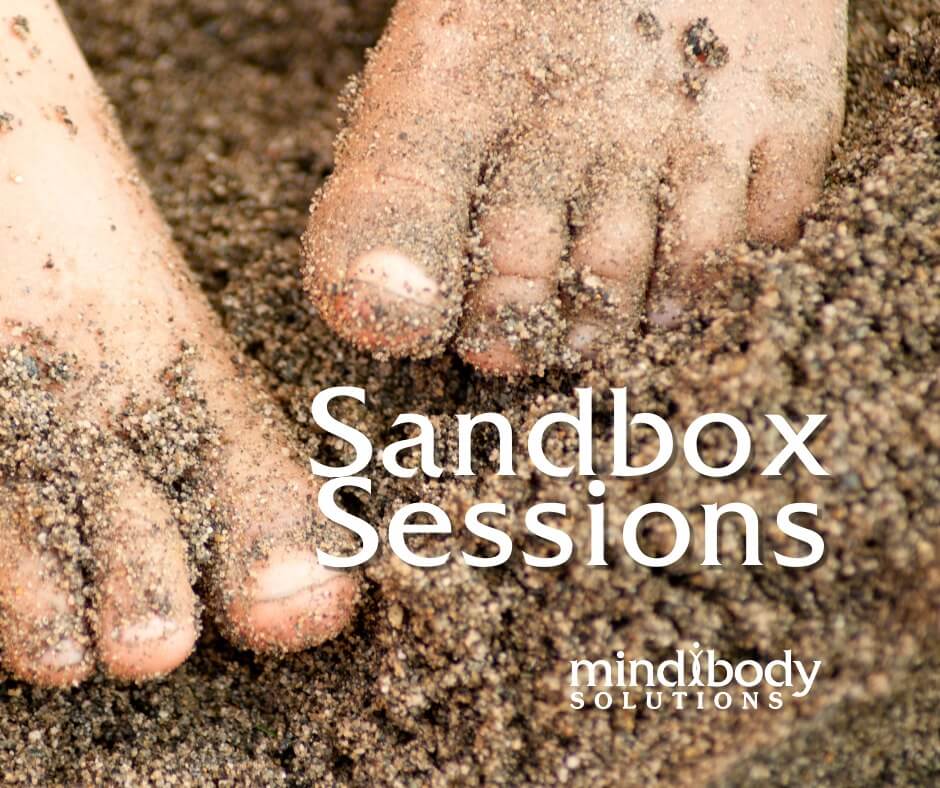 Two feet standing in and covered in sand. Text reading Sandbox Sessions and the Mind Body Solutions Logo at the bottom