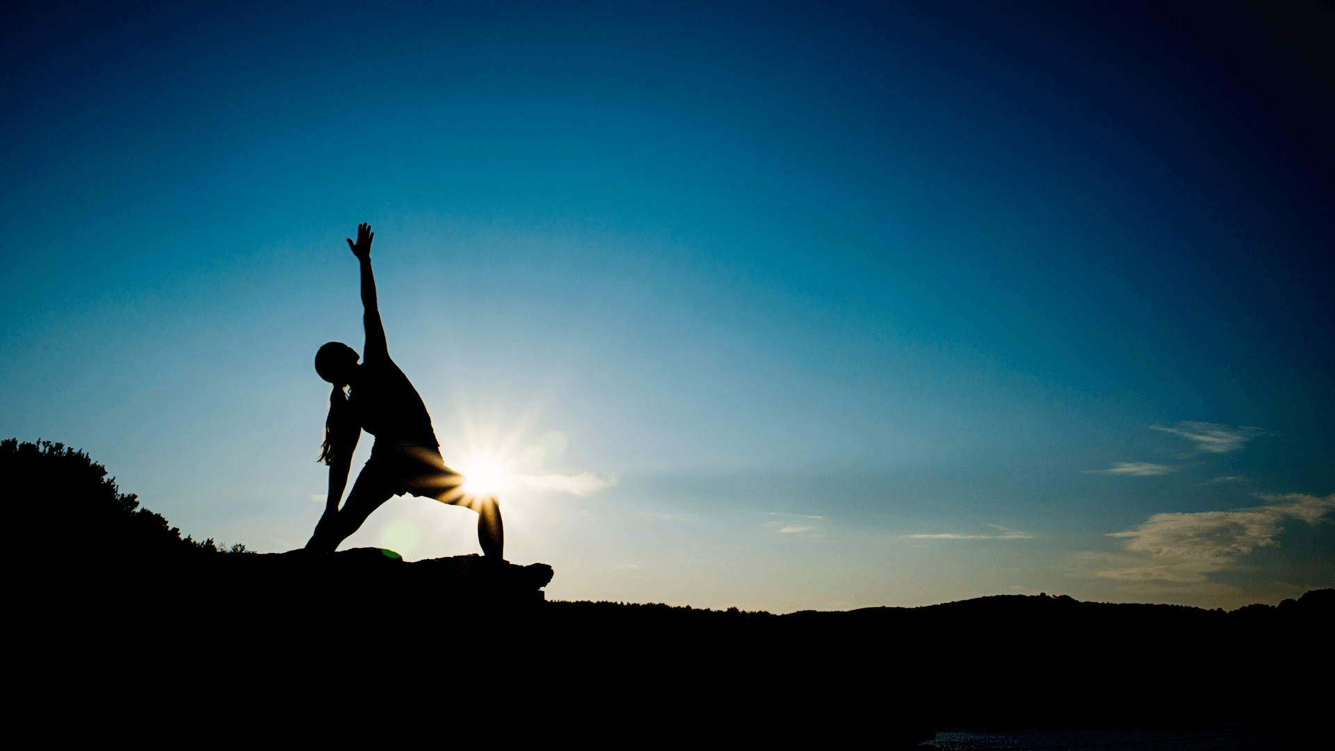 The silhouette of a person standing on a rock ledge, the sun behind them. They are in a Reverse Warrior pose, one hand reaching down an outstretched back leg, the other arm raised to the sky