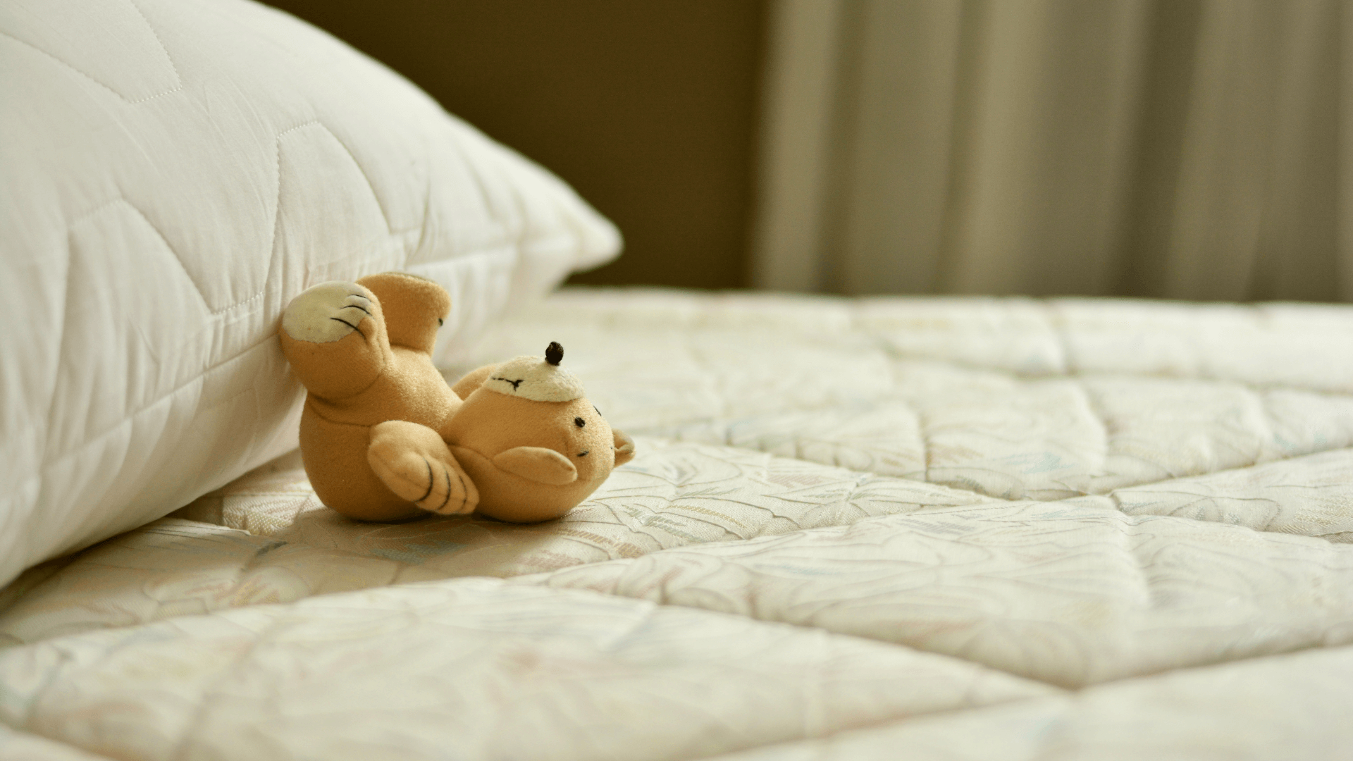 A close up of a bed with cream colored lines. There is a small teddy bear laying on the bed with its head away from the pillow.