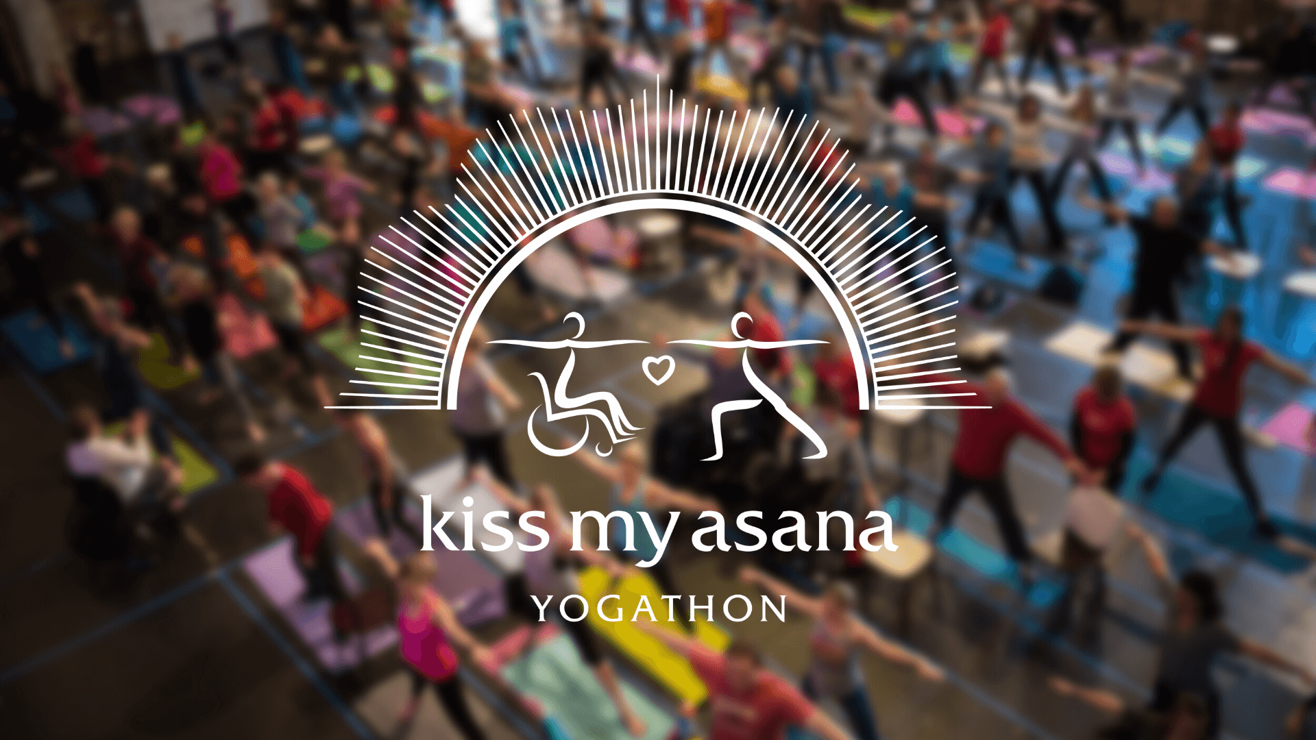 Two stylized figures under an sunburst arch. One figure is in a wheelchair, the other in a Warrior I yoga pose, they are each reaching out to each other and there is a small heart between their hands. Beneath them is text reading "kiss my asana yogathon" The background is a blurred image of an aerial view of many people on a yoga mat with their arms and legs wide.