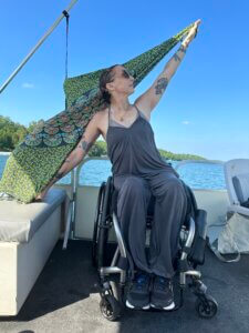  Angelique Lele seated in her manual wheelchair on a boat, wearing a gray jumpsuit, holding a colorful scarf in both hands. She is grounding her hand on a bench next to her and extending the opposite arm.  She is gazing at her extended hand. A view of water behind her. 