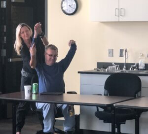 Kevin Kling seated behind a long table, his arms stretched to ceiling. Assistant Molly Bachman is standing behind him, providing reference to his right arm. 