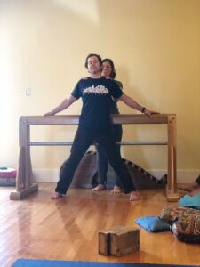 Yoga student Patrick McGuigan standing in front of a large wooden tressler, his arms outstretched and holding the tressler. Teacher trainee Gabrielle Chambliss is behind him providing gentle reference between his shoulder blades. 
