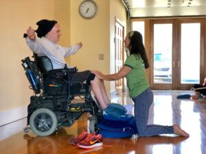 Student Joe Dailey seated in his power wheelchair in a bright sunny yoga studio. His arms are outstretched to shoulder height. MBS instructor Chris Paul is kneeling in front of Joe, her hands on his knees providing gentle reference. They are smiling at each other. 