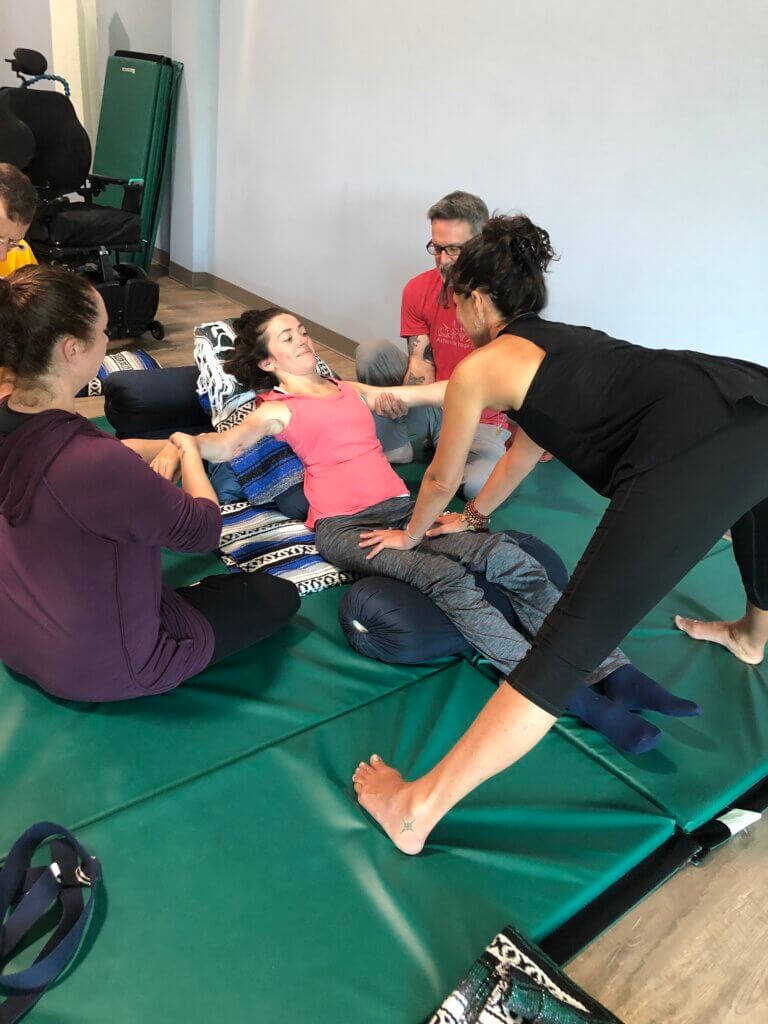 Yoga student Sammie Drost reclined on bolsters, her arms stretched out wide. Three teacher trainees assisting, one supporting each arm and one placing reference on the top of Sammie's thighs. Sammie looks calm and focused. 