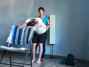 Adaptive student, Rob Ley standing in a right angle, hands on the back of a folding chair in front of him. The chair is on a yoga mat. Sandra Razieli, an adaptive yoga instructor is standing behind him, hands on Rob's hips, providing gentle reference and space. 