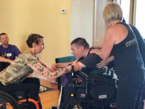 Angelique Lele seated in her manual wheelchair, leaning forward and referencing Chris Becker's arms. Chris is in a power wheelchair and is leaning forward into a downward facing dog pose with reference from Angelique. Two volunteers provide reference to Chris's shoulders and midback. 