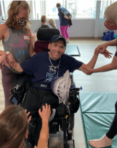 Yoga student Chris Becker seated in his power wheelchair, arms outstretched. Three teacher trainees assisting him, two supporting each arm and one placing reference on his knees. Chris is smiling at the camera. 