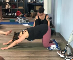 Lindsay Moorehead, a double below the knee amputee, kneeling on a folded yoga blanket. She is in a downward facing dog position, arms outstretched. One of her prosthetic legs is visible propped against the wall next to her. A workshop participant, Jo Mary is kneeling down next to Lindsay. 