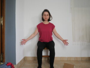 Yoga student Audrey Auzho seated in a chair, feet flat on the floor in front of her. Her arms are stretched to her side, diagonally away from her shoulders. Her eyes are closed. 