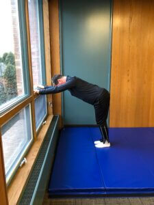 Adaptive student, Jeffrey Riebe, standing on a large blue folding mat, he is in a forward fold, hands on a window at about shoulder height. 