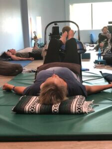 A yoga student laying supine on a large green gym mat, you see her head resting on a folded yoga blanket, her calves resting on a black, armless chair. There is a sandbag resting on her low belly. 