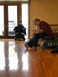 Renee Forst and Sue Fink seated on black folding chairs in a soft yellow yoga studio. They each have one leg extended, a yoga strap around the ball of their foot. They are holding the strap in both hands and gently extending forward. 