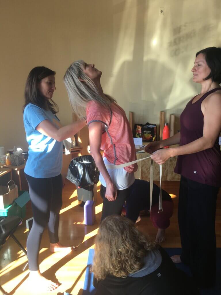 Yoga student Cary Anderson standing in a soft yellow yoga studio. there are two workshop attendees assisting, one standing in front of Cary holding a yoga strap that is around Cary's low back and another supporting Cary's mid back. Cary is in a gentle, supported backbend.