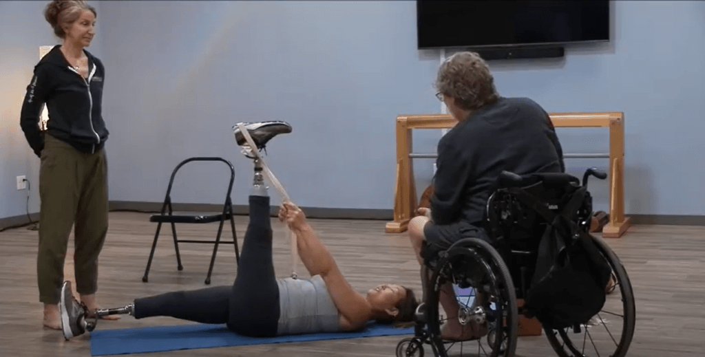 Lindsay Moorehead lying supine on a yoga mat, she is wearing below the knee prosthetics on both legs, Her left leg is raised to the ceiling, holding a strap that is looped around her heel. Amy Samson standing next to her, hands behind her back. Matthew Sanford, in his manual wheelchair is facing away from the camera, toward Lindsay. There is a black folding chair behind them. 
