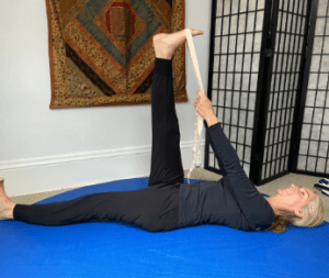 Amy Samson laying supine on a blue yoga mat, her right leg extended to the ceiling. There is a yoga strap looped around her foot and she is holding one side of the strap in each hand. She is smiling. 